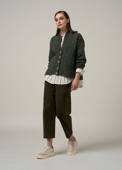Woman wearing a green textured cardigan over a striped shirt with dark pants on a plain background