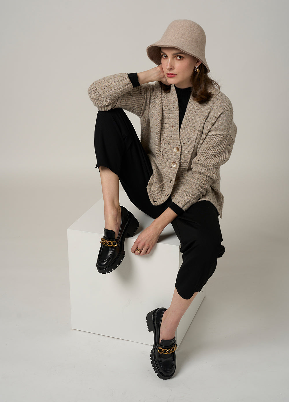 Person wearing a beige cardigan, black pants, and black shoes sitting on a white block against a plain background.